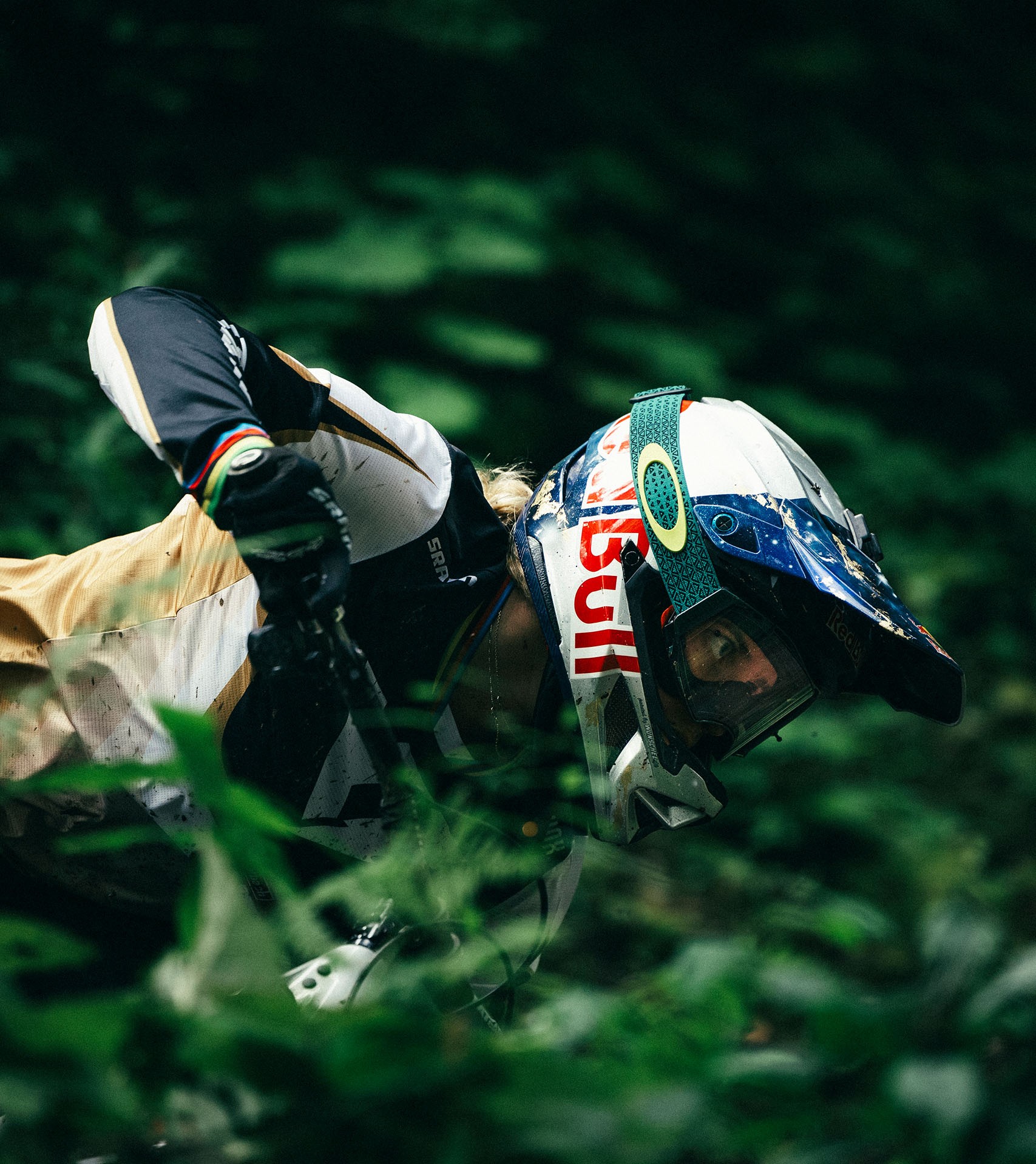 Vali in full gear and protective helmet looks focused as she manoeuvres her bike, framed by lush green foliage.